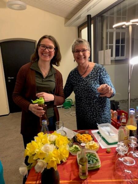 Zwei Frauen stehen hinter einem Tisch, auf dem Zutaten für Cocktails stehen, und lachen in die Kamera