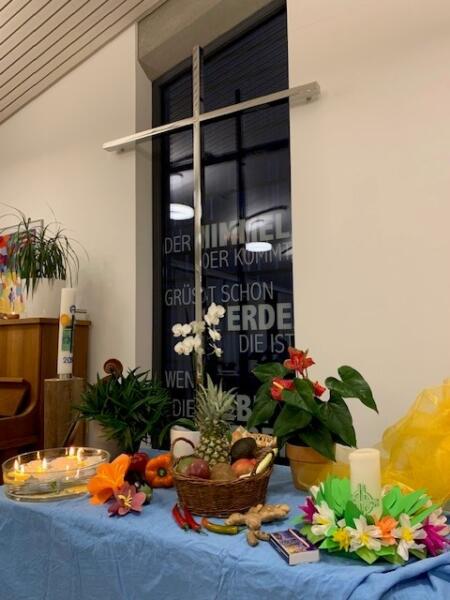 Ein Tisch mit verschiedenen dekorativen Dingen passend zum Weltgebetstag. Links steht eine Wasserschale mit Teelichtern in der Mitte steht ein Weidenkorb mit Früchten und rechts eine Kerze um die ein Blumenkranz gelegt ist.